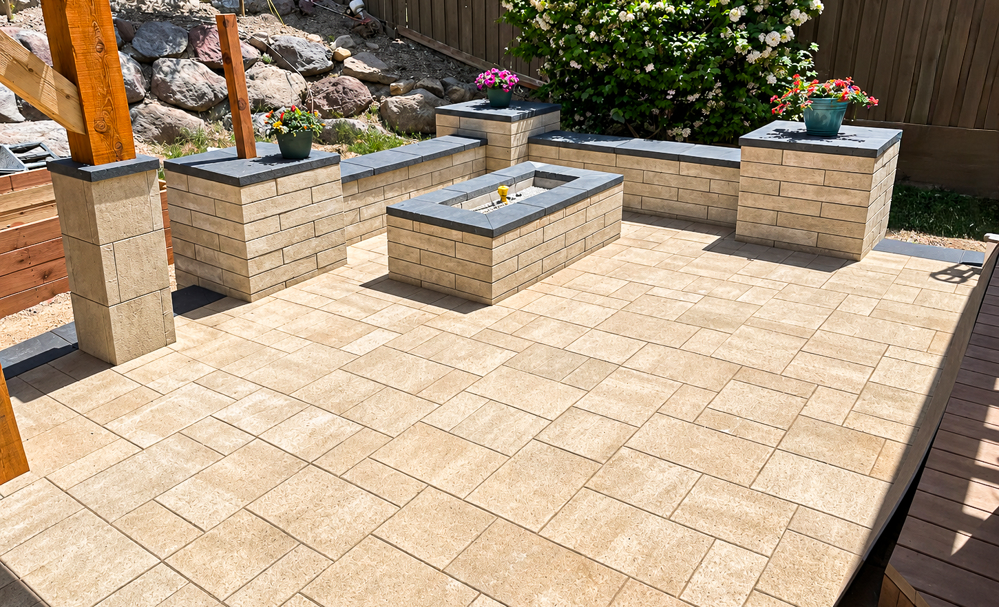 Tan paver backyard patio with a matching stone fire pit, low retaining walls and potted flowers, custom paver patios by Empire Pavers in Utah