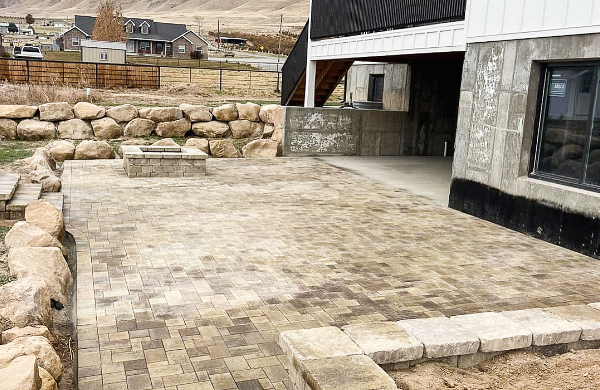 Tan paver patio with a square stone fire pit and boulder retaining wall behind a Utah home, retaining wall project in Morgan, UT