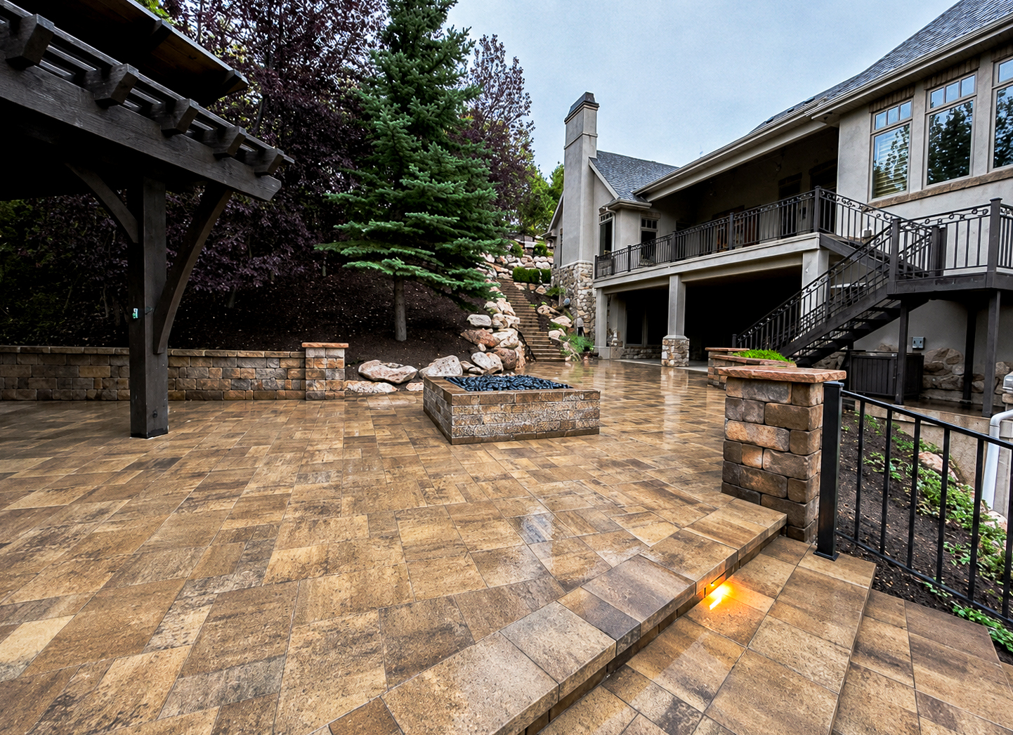 Multi-level paver patio at dusk with a square fire pit, stone pillars and a wood pergola, retaining walls by Empire Pavers in Utah